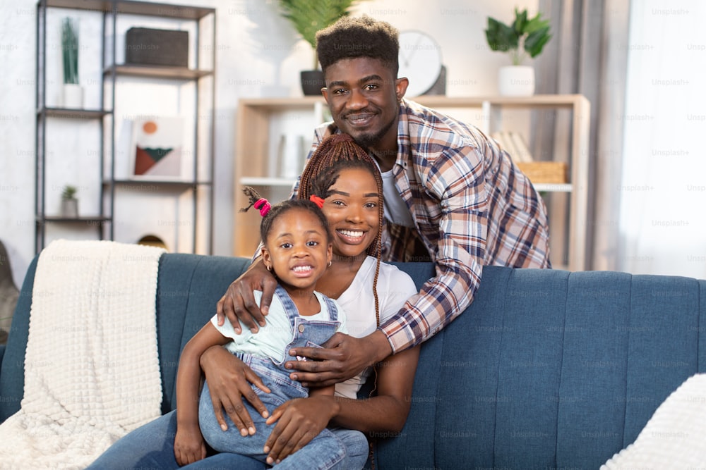 Ein gutaussehender afroamerikanischer Mann, der seine charmante Frau mit hübscher Tochter umarmt, während er auf einer bequemen Couch sitzt. Glückliche Familie, Liebe und Erziehungskonzept.