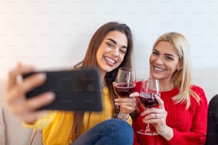 Laughing woman friends hugging each other on sofa while taking selfie photo on smart phone. Lovable blond and brunette girls expressing positive emotions to camera while drinking wine