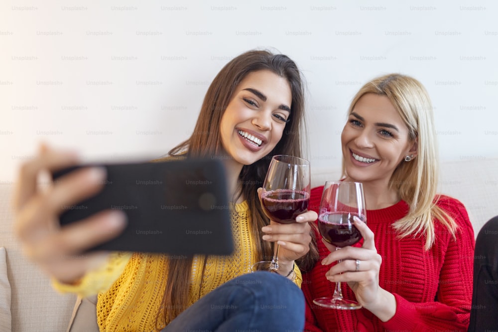 Amiche donne che ridono che si abbracciano sul divano mentre scattano foto selfie sullo smartphone. Adorabili ragazze bionde e brune che esprimono emozioni positive alla telecamera mentre bevono vino