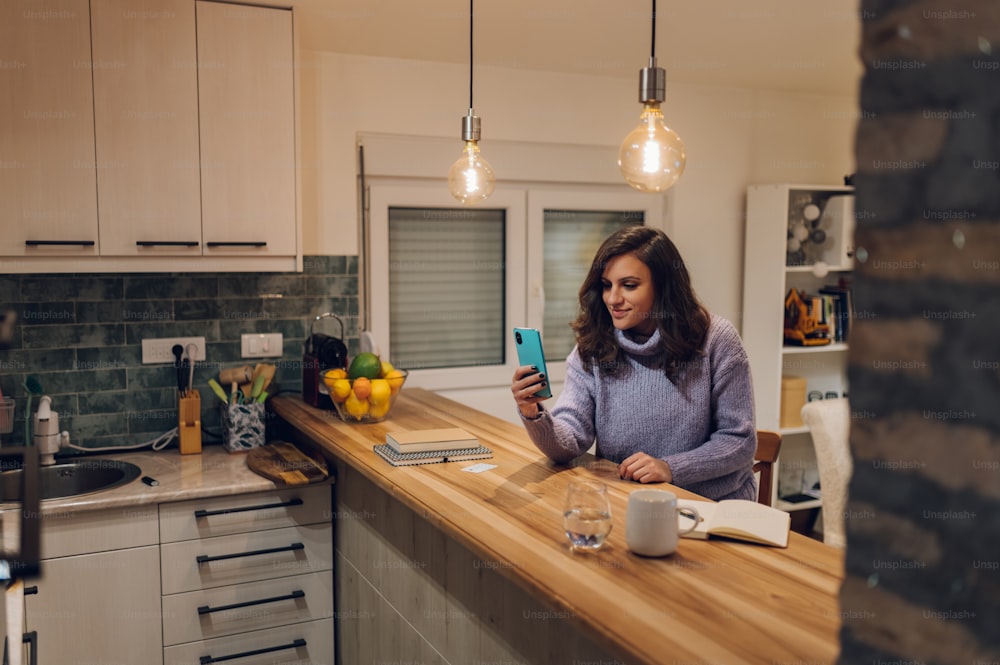 Young hispanic woman is using smartphone for a video call to stay in touch with family while in a kitchen at home. Enjoying distance communication. Digital life concept.