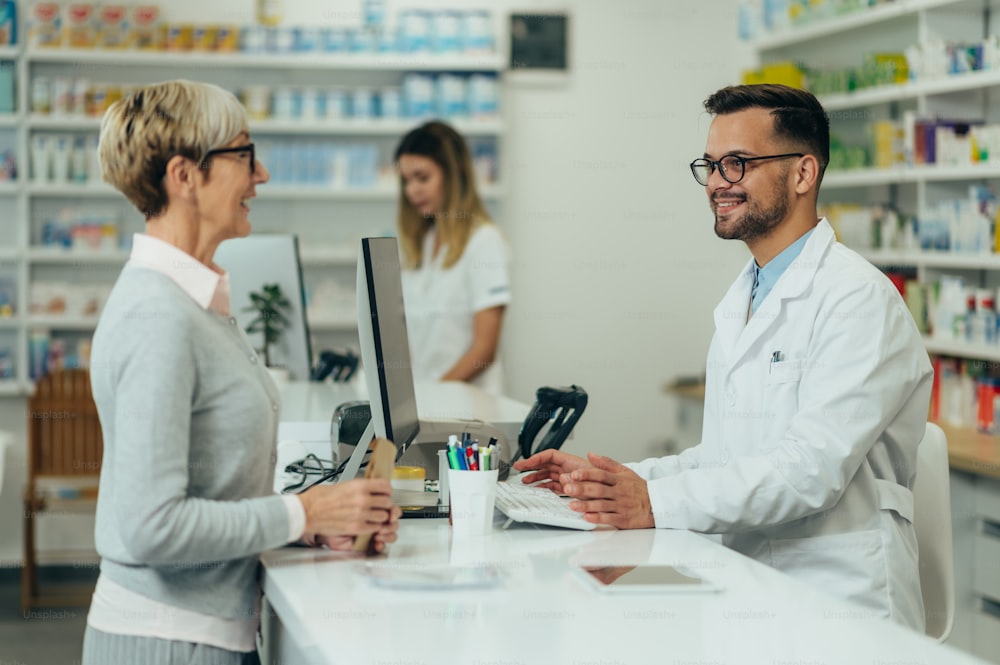 Junger männlicher Apotheker gibt verschreibungspflichtige Medikamente an ältere weibliche Kunden in einer Apotheke mit Apothekerin im Hintergrund