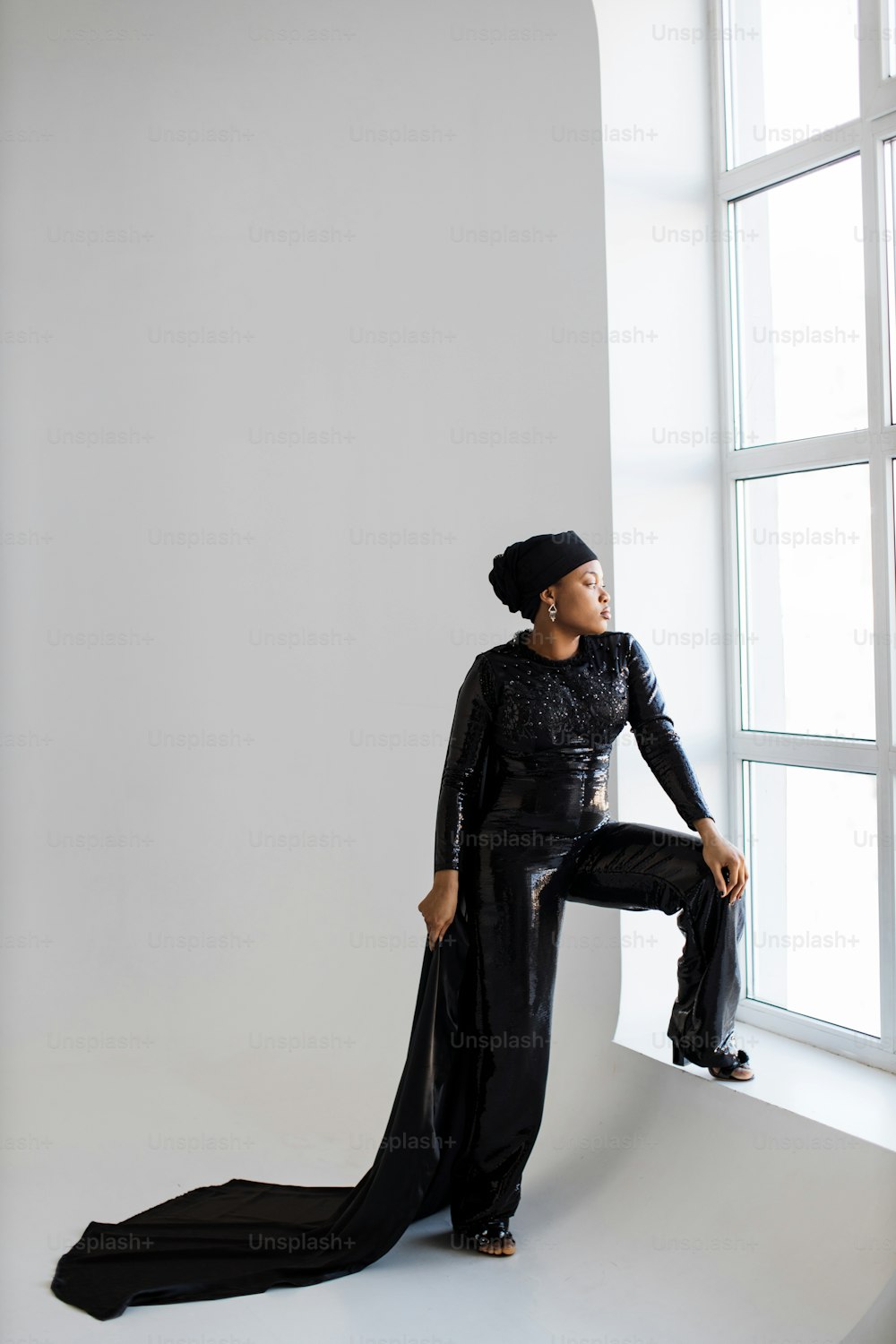 Gorgeous african lady wearing black trendy hat and dress with long cape looking at window while standing at white studio. Confidence and beauty of mature woman.