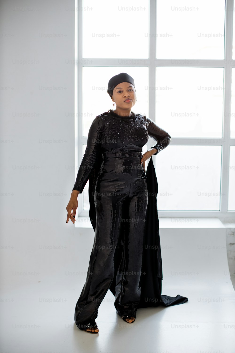 Elegante mujer afroamericana islámica con traje de noche negro y sombrero mirando a la cámara mientras posa sobre fondo blanco. Concepto de belleza, moda y elegancia.