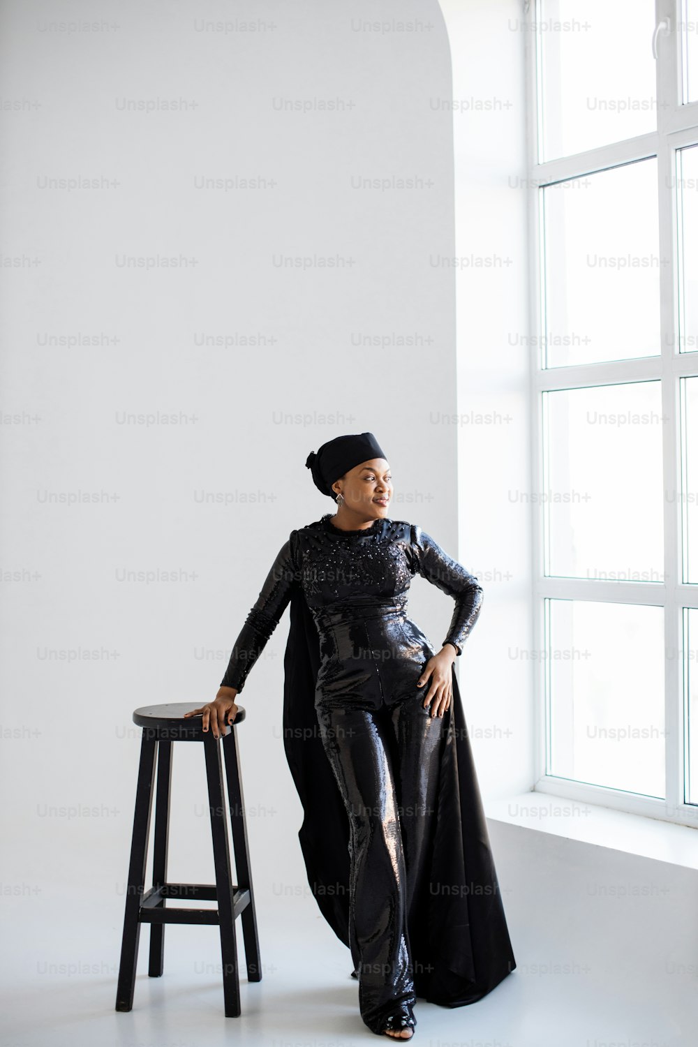 Atemberaubende afroamerikanische Frau mit stilvollem schwarzen Hut und Kleidung, die sich auf einen Hochstuhl stützt und zum Fenster schaut. Weißer Studiohintergrund.