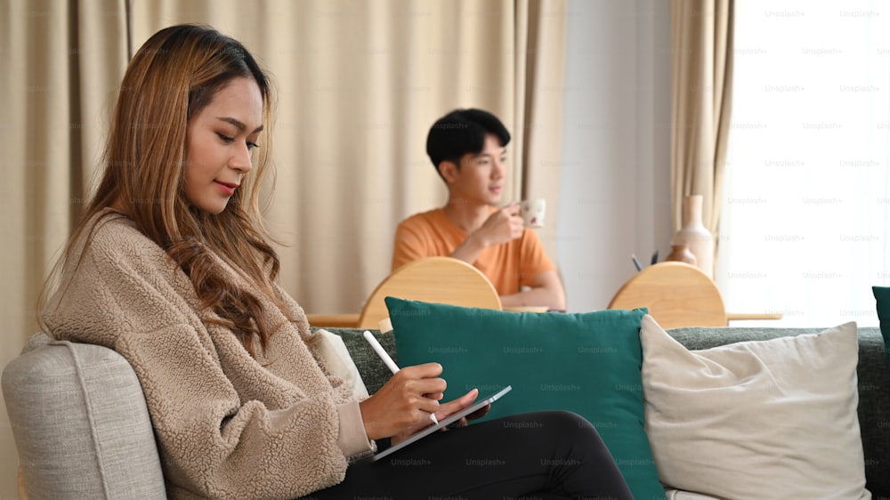 소파에서 디지털 태블릿을 사용하는 젊은 아시아 여성과 그녀의 남편이 배경에 앉아 있다.