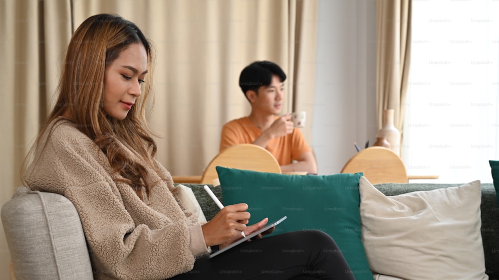 Young asian woman using digital tablet on sofa and her husband sitting in background.