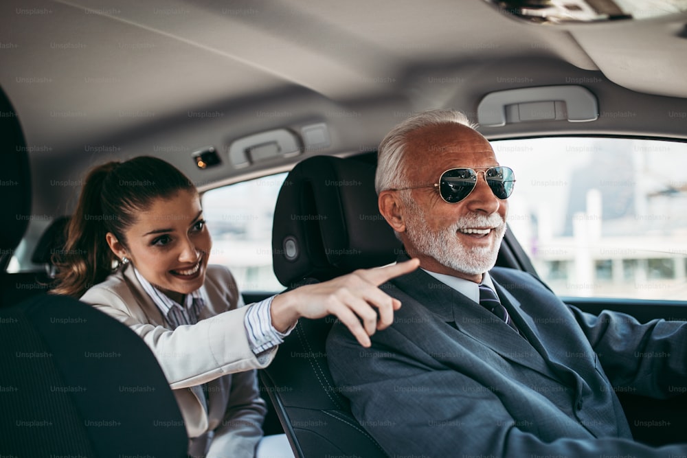 Beau conducteur senior et jeune femme d’affaires assis sur la banquette arrière dans une voiture de luxe. Ils parlent et sourient. Transport dans le concept d’entreprise d’entreprise.