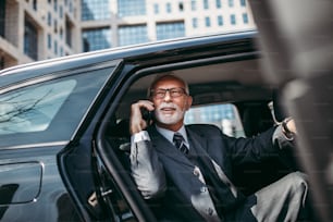 Hombre de negocios senior de buen aspecto sentado en el asiento trasero en un automóvil de lujo. Abre las puertas del coche y sale o sale. Gran negocio que se construye en el fondo. El transporte en el concepto de negocio corporativo.
