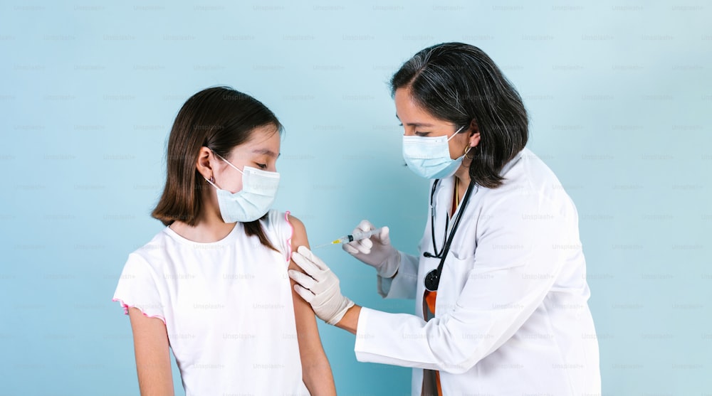 Latin Medical Frau Ärztin oder Krankenschwester gibt Coronavirus-Impfstoff Schuss zu hispanischem Kind Mädchen über blauen Studiohintergrund