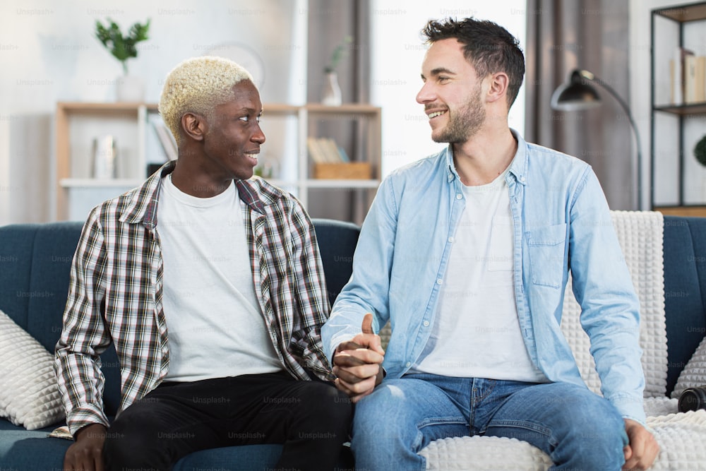 Happy caucasian and african american men holding hands and looking on each other while sitting on couch. Two loving people in casual attire spending free time together at home.