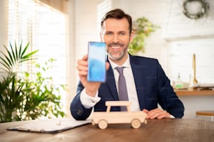 Hombre de negocios, concesionario de automóviles sosteniendo un teléfono móvil, mostrando una pantalla vacía a la cámara. La maqueta del coche de madera sobre el escritorio. Detalles del seguro, alquiler de coches. Concepto de vehículo seguro.