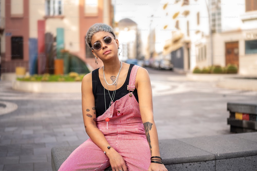 Jeune fille moderne à la mode aux cheveux courts portant des lunettes de soleil et beaucoup de bijoux. Style de rue de la ville. Photo en plein air.