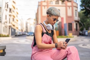 携帯電話を使って短い髪型をし、ソーシャルメディアをスクロールし、街の通りに座っているファッショナブルなモダンな若い女性。