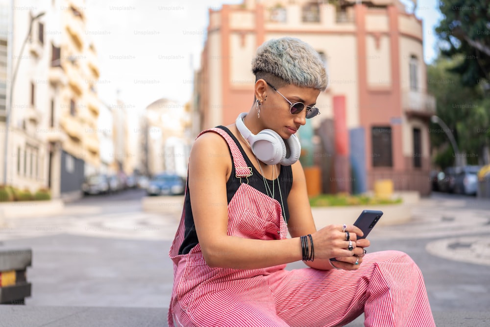 짧은 헤어스타일을 한 세련된 현대 젊은 여성은 휴대폰을 사용하여 소셜 미디어를 스크롤하고 도시 거리에 앉아 있다.