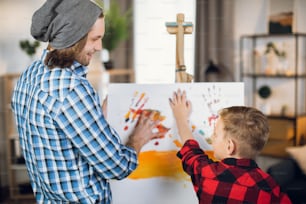 Padre caucásico cariñoso enseñando a su pequeño hijo a dibujar sobre lienzo. Hombre guapo y chico lindo con camisas a cuadros usando pinturas de colores en casa.