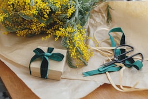 Bonne fête de la femme et bonne fête des mères. Bouquet de mimosa élégant et boîte-cadeau sur fond en bois. Belles fleurs de mimosa en papier kraft et cadeau avec ruban vert sur le rebord de la fenêtre. 8 mars