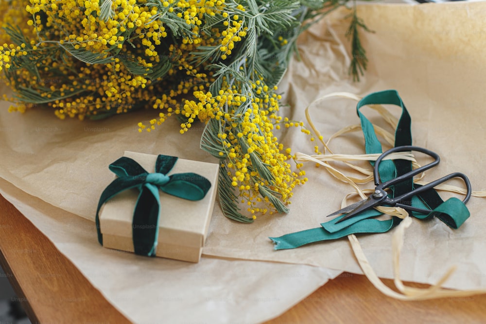 Happy womens day and Happy Mothers day. Stylish mimosa bouquet and gift box on wooden background. Beautiful mimosa flowers in craft paper and gift with green ribbon on windowsill. 8 march