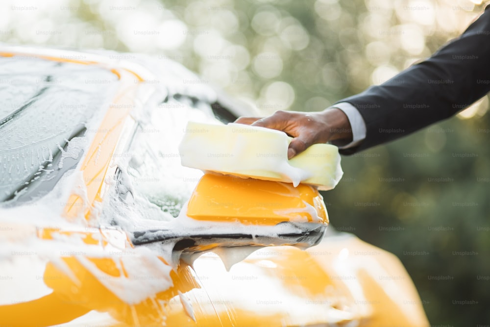 Image recadrée en gros plan de la main d’un jeune homme d’affaires africain avec une éponge jaune lavant le rétroviseur de sa voiture dans un lave-auto en libre-service à l’extérieur. Voiture de luxe recouverte de mousse.
