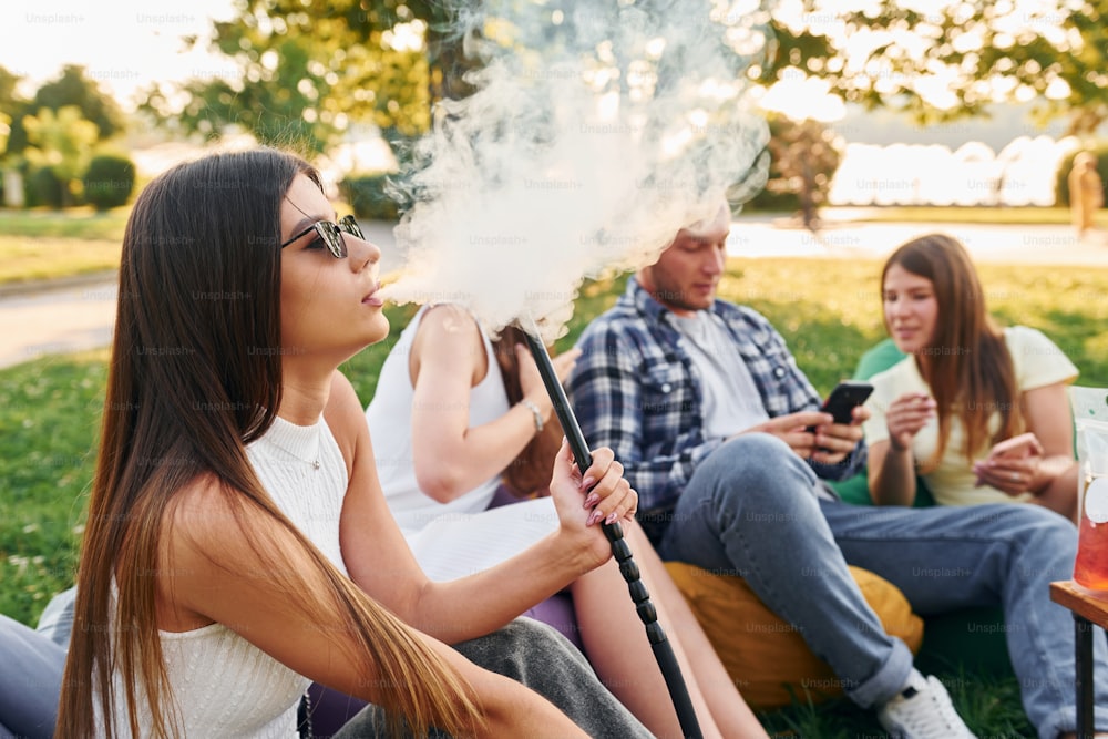 Sentado nos sacos da poltrona e fumando narguilé. Grupo de jovens tem uma festa no parque durante o dia de verão.