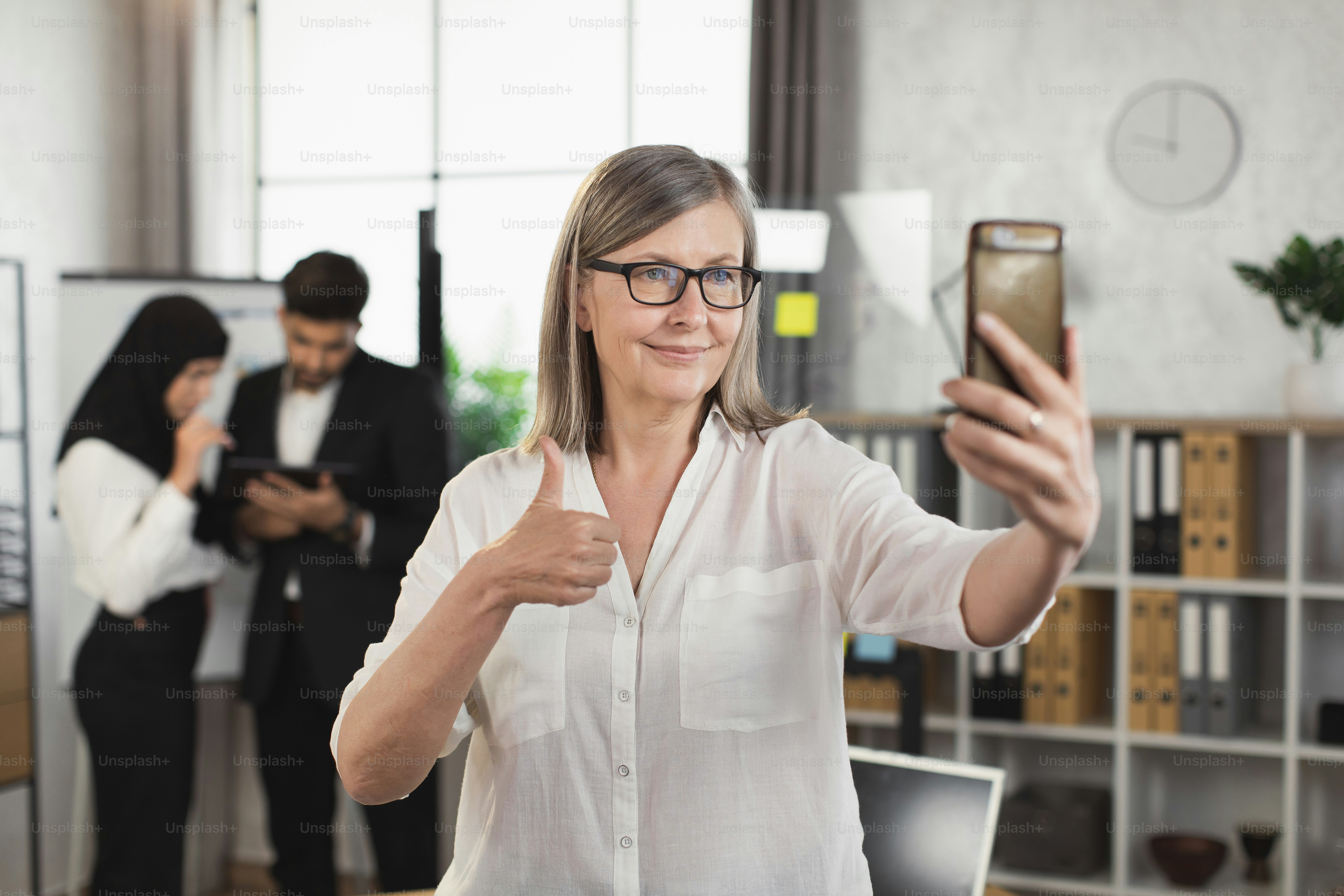 Dama de negocios madura segura de sí misma con anteojos que usa un teléfono inteligente para videoconferencia en una oficina moderna y muestra el pulgar hacia arriba. Desenfoque el fondo de dos colegas multirraciales usando una tableta digital.