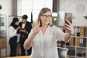 Confident mature business lady in eyeglasses using smartphone for video conference at modern office. Blur background of two multiracial colleagues using digital tablet.