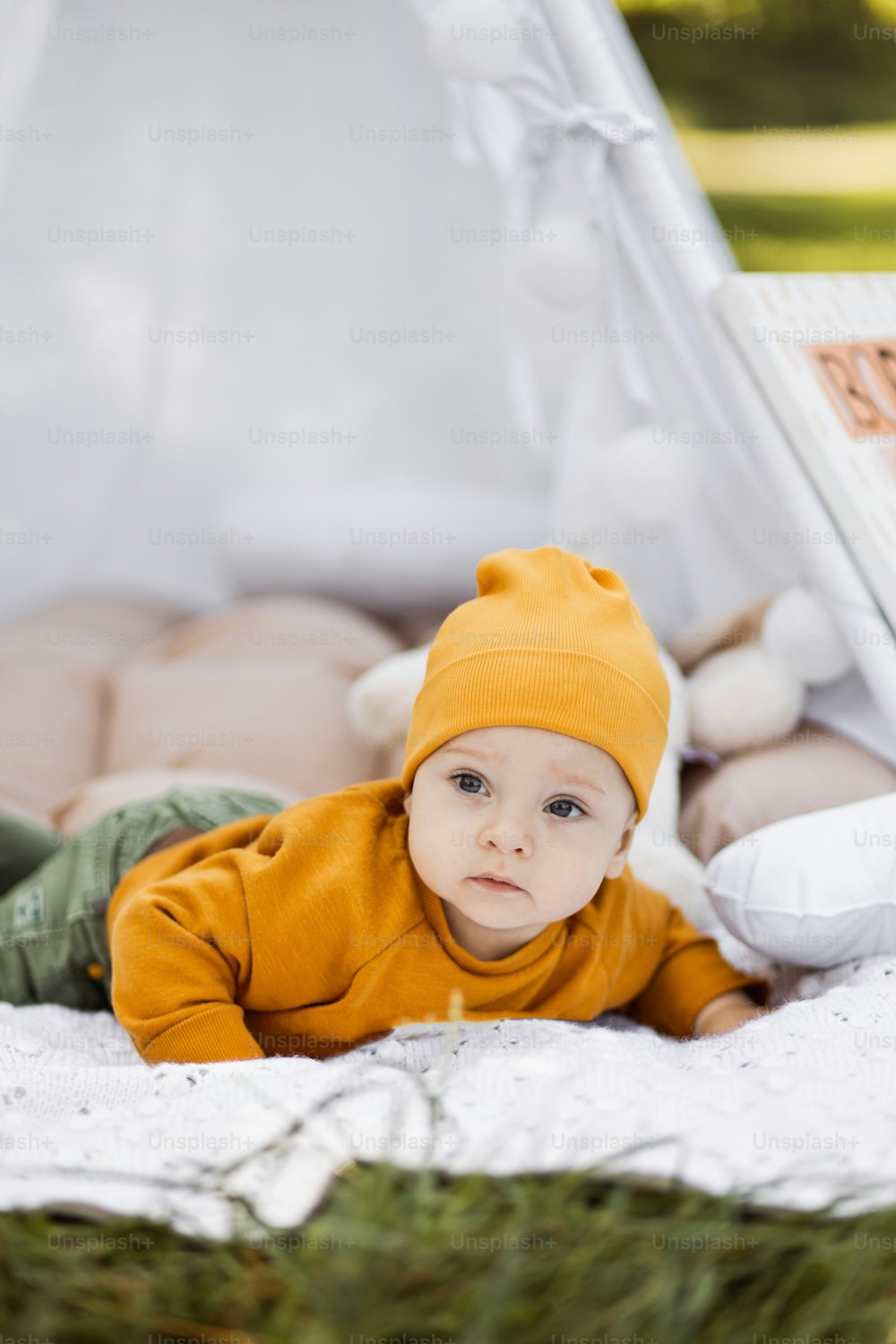 Portrait of cute caucasian baby boy in stylish wear and hat lying in colorful wigwam with various toys around. Pretty toddler on picnic among green summer nature.