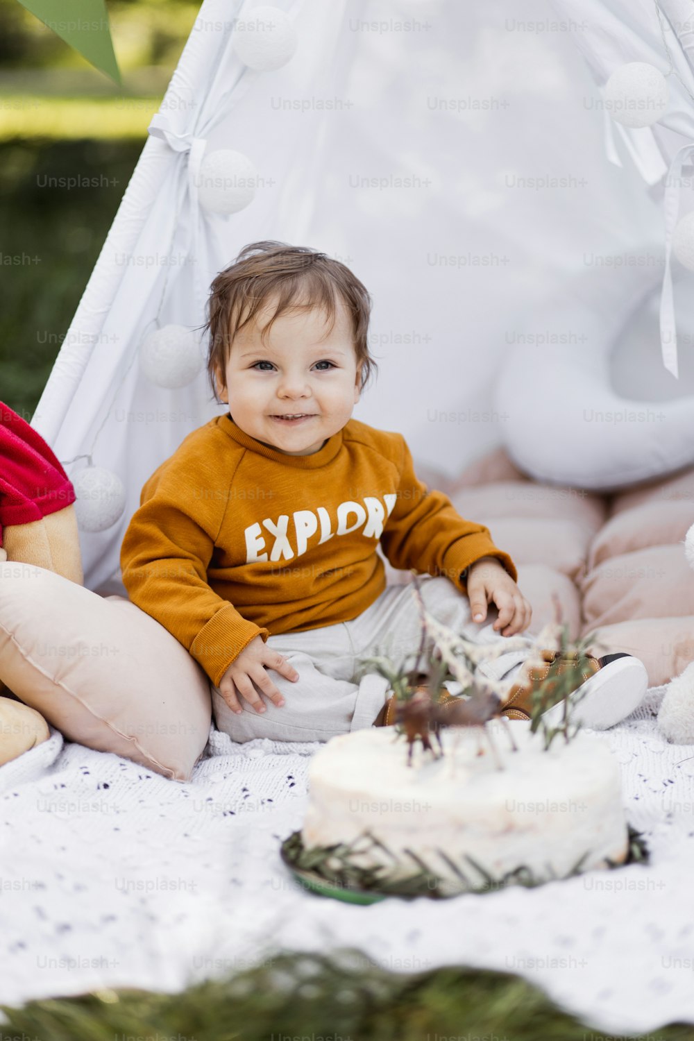 Pretty little baby boy in casual wear looking on beautiful sweet cake while sitting in toy wigwam. Birthday celebration with colorful decoration.