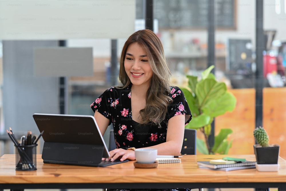 コーヒーショップに座ってコンピュータータブレットワイルとオンラインで働いている幸せな若いアジアの女性。