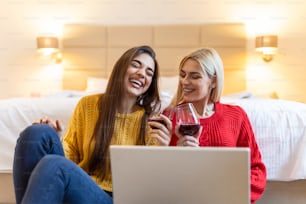 Two attractive girls, cheerful best friends having fun,watching movies on laptop and drinking red wine at home. Two glasses of white wine in hands
