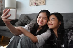 Two Asian girls taking selfie with smart smartphone while sitting together in living room.