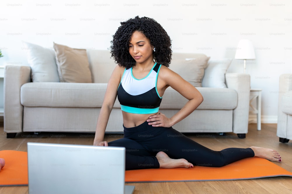 Une femme africaine adulte fait des exercices de yoga et de musculation sur un tapis dans son salon. Elle suit une vidéo de cours d’exercices en ligne sur son ordinateur portable.