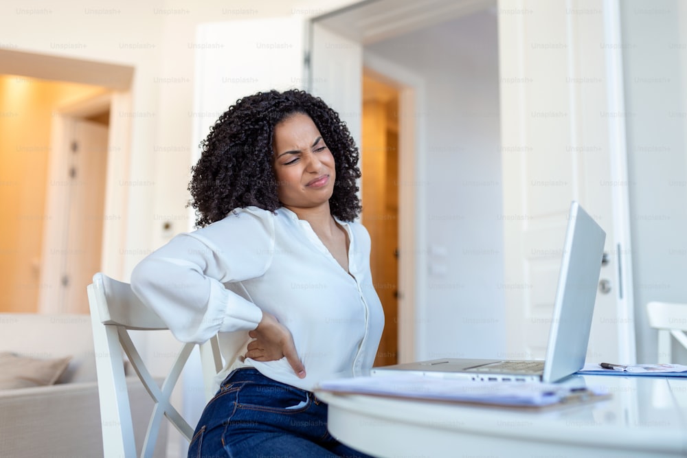 Young businesswoman having back pain while sitting at office desk. Businesswoman Holding Her Back While Working On Laptop At Office Desk