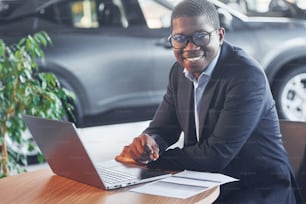 Office worker is sitting in autosalon by table with laptop.