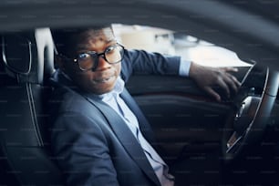 In glasses. Young african american businessman in black suit is in the automobile.