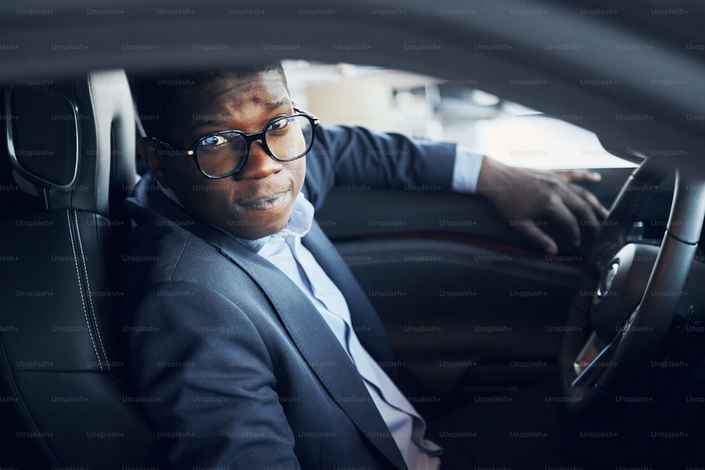 In glasses. Young african american businessman in black suit is in the automobile.