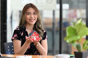 Happy young woman holding small house model and smiling at camera. Real estate investment concept.