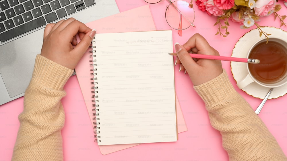 宿題をしながら、美しいピンクの机で学校のノートにエッセイを書いている女子大生。上面図、フォーカスハンド