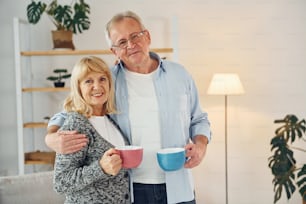 Stehen und Tassen mit Getränken halten. Älterer Mann und Frau sind zusammen zu Hause.