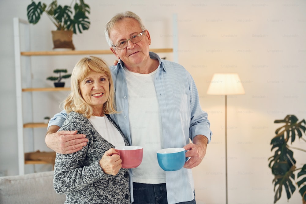 Standing and holding cups of drinks. Senior man and woman is together at home.