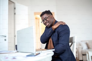 自宅でパソコンで仕事をしているアフリカのビジネスマンは、肩に痛みを感じています。肩や背中の痛みに悩まされている男性は、自宅でノートパソコンを使って座って仕事をしています。