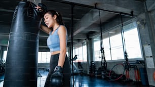 Mulher atlética asiática forte em roupas esportivas usando luvas de boxe fazer exercício de treino socando saco de boxe na academia de fitness. Saudável ajuste e firme feminino fazer treinamento esportivo e musculação no clube esportivo.