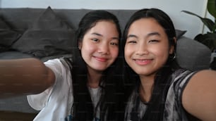 Fröhliche asiatische Mädchen, die in die Kamera schauen, machen Selfies, während sie zusammen im Wohnzimmer sitzen.