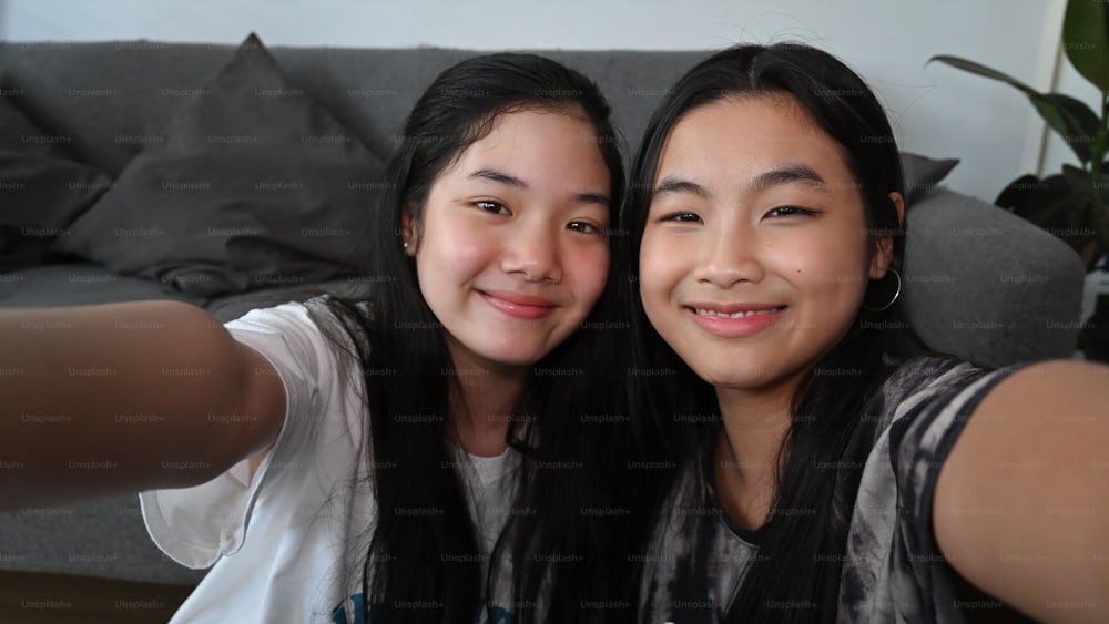 Joyful Asian girls looking at camera make selfie while sitting together in living room.