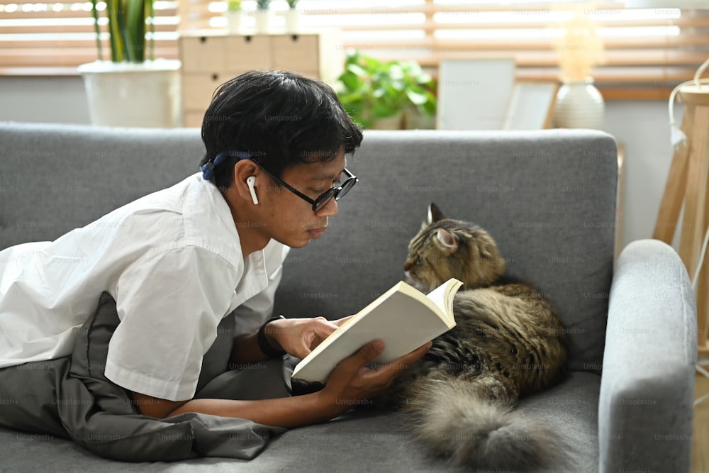 캐주얼한 남자는 사랑스러운 고양이와 함께 소파에 누워 책을 읽고 있다.