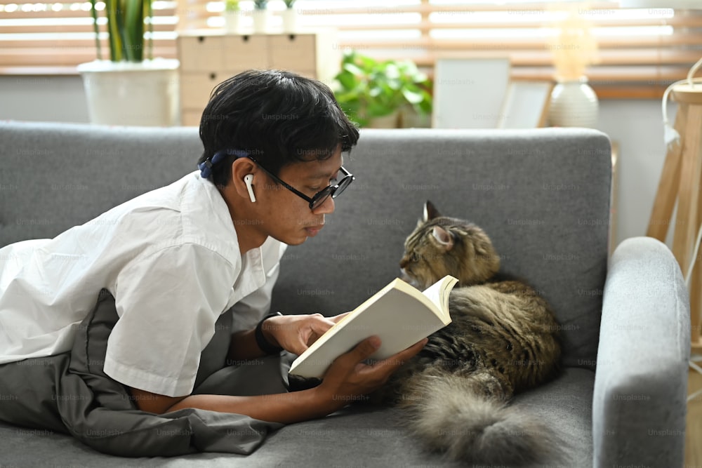 Casual man lying on couch with his lovely cat and reading book.