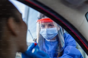 PPE、N95マスク、フェイスシールド、個人用保護ガウンを着用した医師または看護師が、車/道路のそばに立ってCovid-19ウイルスのスクリーニング、鼻腔スワブテストを行います。
