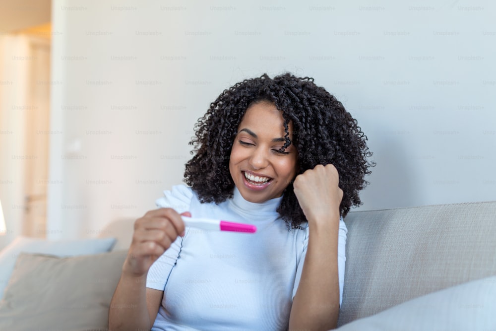 幸せそうに妊娠検査を見ている若い女性。ついに妊娠。妊娠検査を見て、自宅のソファに座りながら微笑む魅力的な黒人女性