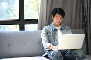 自宅のソファで休んで、コンピューターのラップトップでインターネットを閲覧するカジュアルなアジア人男性。