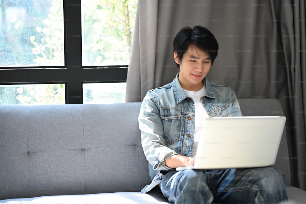 Uomo asiatico casuale che riposa sul divano a casa e naviga in internet con il computer portatile.