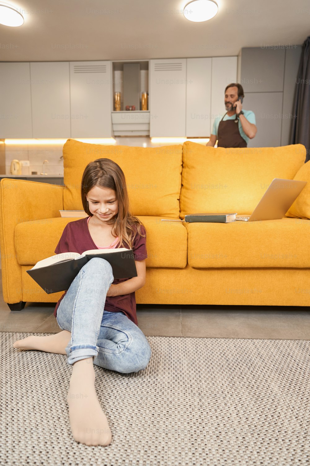 Smiling teenager looking at the book on the floor while her father talking on a mobile on the background